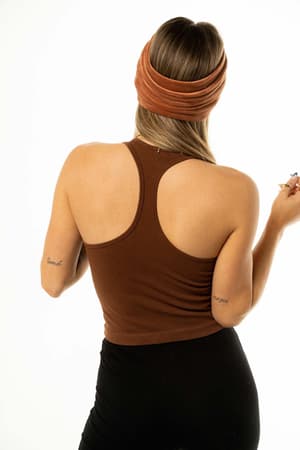 A woman with long hair, wearing the High Neck Racerback Brami in brown and black pants, stands facing away from the camera. She has small arm tattoos and holds a small object in her right hand.