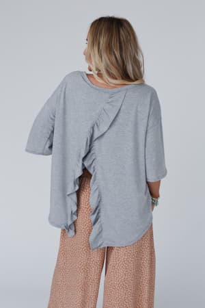 A woman with light brown hair, seen from behind, wears The Nest Must Have High Low Tee in heather gray with wide sleeves and a diagonal ruffle across the back, paired with flowy peach boho patterned pants.