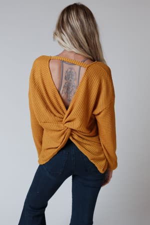 A woman with long blonde hair wears The Nest Flattering Open Back Thermal Knit Top in mustard with a waffle knit and twisted back, showing a large tattoo. She styles this boho look with dark blue jeans.