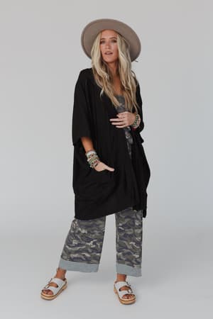 A woman in a wide-brimmed hat, camo print pants, white sandals, colorful bracelets, and The Nest Boho Essential Hooded Ribbed Sweater - Black stands against a plain background, looking ahead with confidence.