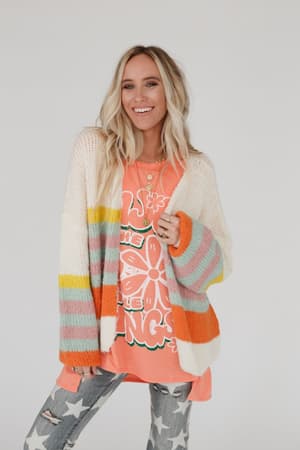 A smiling woman with long blonde hair wears the Retro Babe Striped Cardigan - Cream over a bright orange graphic tee and star-print gray jeans, standing against a plain light background.