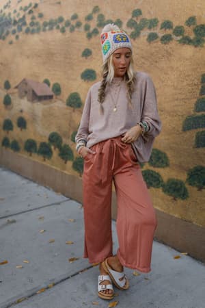 A woman wearing The Nest Fly With Me Batwing Sleeve Knit Sweater in Sand, colorful beanie, and wide-legged salmon pants stands on a sidewalk near a mural of hills and a barn. She completes her look with sandals and layered necklaces.