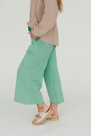 A person stands sideways with one hand in their pocket, wearing the Lucky One Wide Leg Pant in green, a beige long-sleeve top, and beige thick-soled sandals with buckled straps. The background is plain white.
