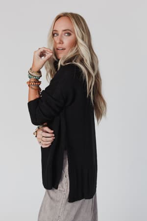 A woman with long blonde hair wears the loose-fitting black Danny Sweater and light pants, accessorized with colorful beaded bracelets, as she poses against a plain light background, glancing over her shoulder.