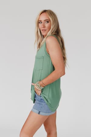 A woman with long blonde hair wears The Nest Shelli Scoop Neck Tank Top in Sage, styled in a relaxed fit with denim shorts. She stands sideways with a hand in her pocket against a plain light background, accessorized with several bracelets.