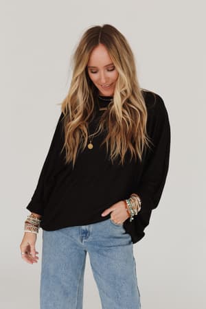 A woman with long, wavy blonde hair smiles while wearing the Fly With Me Batwing Sleeve Knit Sweater in black, paired with light blue jeans, layered necklaces, and colorful beaded bracelets against a plain light background.