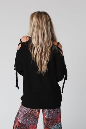 A woman with long, wavy blonde hair stands with her back to the camera, wearing the Roxy Criss Cross Tie Shoulder Sweater in black and colorful patterned pants. The plain background is light-colored.