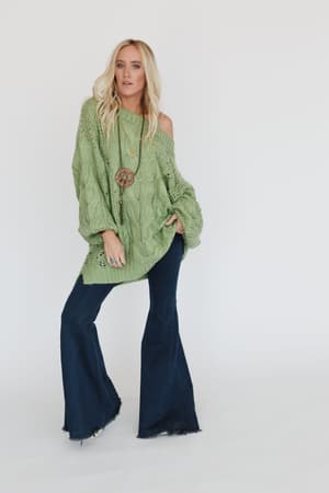 A woman with long blonde hair wears the Golden Hour Bubble Sleeve Sweater Dress in light green, styled off one shoulder, paired with dark blue flared jeans and a long necklace, standing against a plain white background.