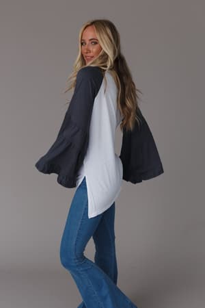 A woman with long blonde hair poses sideways, wearing the Bell Sleeve Baseball Tee in charcoal and blue flared jeans, standing against a plain grey background.