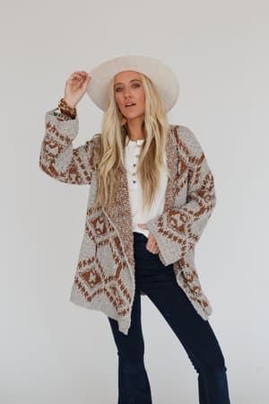 A woman with long blonde hair wears the Free to Roam Cardigan - Gray over a white shirt, paired with dark jeans and a white hat. She stands against a plain light background, holding the brim of her hat with one hand.