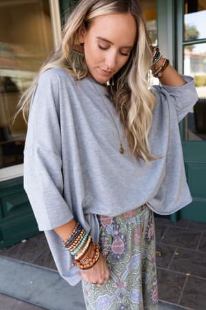 A woman with long blonde hair wears The Nest Cozy Cool Oversized Tee in Heather Gray with patterned pants and layered jewelry, standing outside by a green door and window.