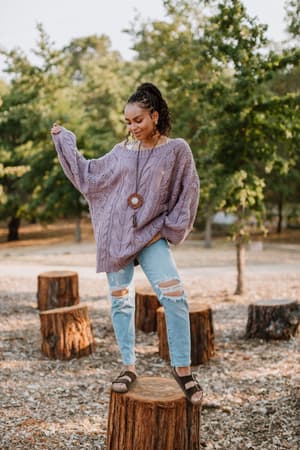 A woman stands confidently on a tree stump in a park, surrounded by cut logs and greenery, wearing the Golden Hour Bubble Sleeve Sweater Dress in Lavender.
