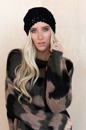 A woman with long blonde hair wears the Wild Wanderer Sweater - Camo and a black knit beanie, standing in front of a light wood background and looking at the camera with a relaxed expression.