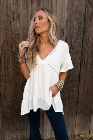 A woman with long blonde hair poses against a wooden wall, wearing the More Than Meets The Eye Tunic Top in ivory, paired with dark jeans and colorful bracelets. One hand is in her pocket as she looks to the side.