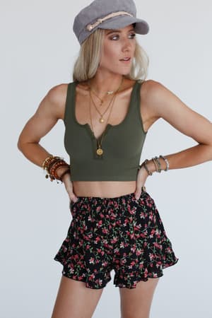 A woman, hands on hips, looks to the side wearing an olive Hook It Up Ribbed Brami, black floral ruffled shorts, a gray cap, layered necklaces, and multiple bracelets on both wrists.