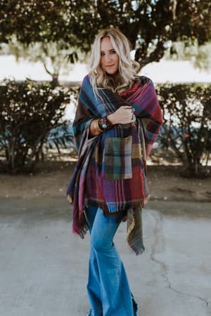 A woman with wavy blond hair stands outdoors in the Cozy Plaid Pocket Ruana Wrap - Fuchsia, paired with blue flared jeans and bracelets, surrounded by trees and bushes.