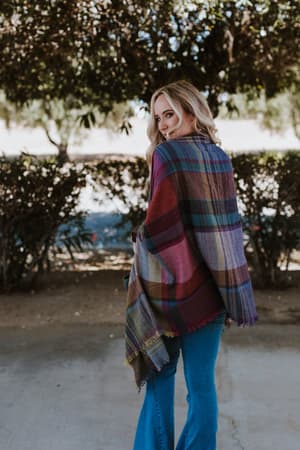 A woman with blonde hair stands outdoors, smiling over her shoulder. She is wearing a Cozy Plaid Pocket Ruana Wrap - Fuchsia and blue jeans, surrounded by trees and bushes.