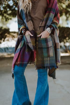 A woman stands outdoors with her hands in the pockets of a Cozy Plaid Pocket Ruana Wrap - Fuchsia, paired with a brown top, blue flared jeans, and bracelets. Only her body from the shoulders down is shown.
