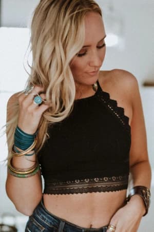 Wearing the Light Of The Moon Crochet Trim Bralette in black, a woman with long blonde hair pairs it with blue jeans and layered jewelry, looking down as she holds a strand of her hair.
