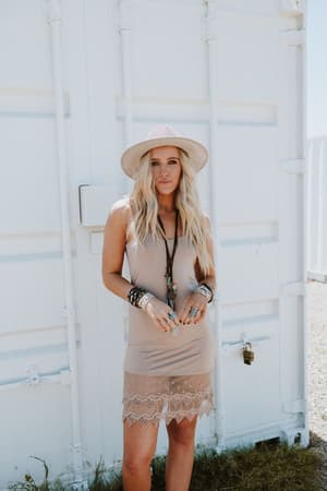 A woman with long blonde hair wears the Layering Love Lace Slip in mocha and a wide-brimmed hat. She stands outdoors before a white metal container on a sunny day, gazing at the camera and accessorized with multiple bracelets.