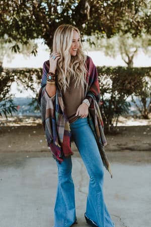 A woman with long blonde hair, wearing a brown top, blue flared jeans, and the Cozy Plaid Pocket Ruana Wrap - Fuchsia stands smiling outdoors on a sidewalk with trees and bushes in the background.