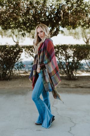 A woman in flared blue jeans and a vibrant Cozy Plaid Pocket Ruana Wrap - Fuchsia stands outdoors on a concrete path, with trees and bushes in the background.