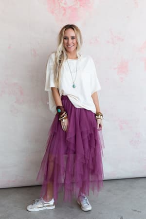 A smiling woman with long blonde hair stands by a light pink wall, wearing a white loose t-shirt, the mauve Boho Ballerina Skirt, white sneakers, and layered bracelets and necklaces for a relaxed, cheerful look.