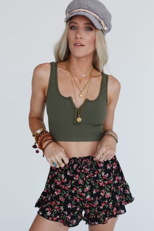 A woman wears the Hook It Up Ribbed Brami in olive, styled with layered necklaces, bracelets, rings, a gray hat, and black floral ruffle shorts against a plain white background.