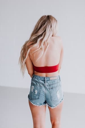 A woman with long, wavy blonde hair faces away, wearing the Baroque High Neck Velvet Bralette in wine and distressed denim shorts against a plain white background.