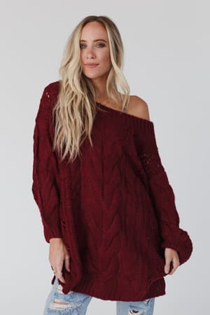 A woman with long blonde hair wears the Golden Hour Bubble Sleeve Sweater Dress in burgundy, styled off-the-shoulder, standing against a plain light background.