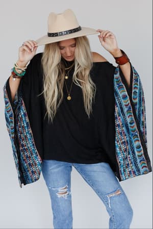 A woman in a beige hat, layered necklaces, bracelets, ripped blue jeans, and the Fallon Embroidered Sleeve Poncho - Black poses against a plain background, holding her hat and looking down.
