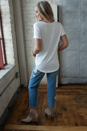 A woman stands indoors facing away, wearing a white Scoop Neck Bralette Tee, ripped blue jeans, and beige fringe ankle boots on a wooden floor near a window and exposed brick wall.