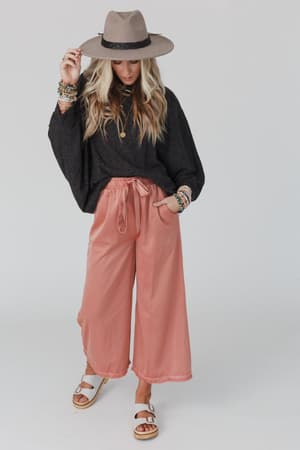 A woman wears The Nest Fly With Me Batwing Sleeve Knit Sweater in charcoal, paired with pink wide-leg pants, bracelets, and white sandals, standing against a white background and looking down while holding her hat brim.