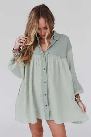 A woman with long, wavy blonde hair wears the Sunday Kind Of Love Dress in sage. The oversized boho dress has long sleeves. She looks down, holding her hair, accessorized with bracelets and rings against a plain white background.
