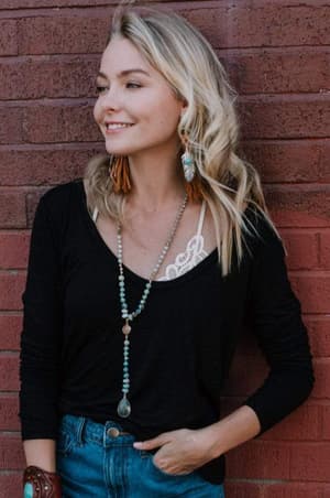 A smiling woman with long blonde hair stands against a brick wall, wearing the Ryder Long Sleeve Bralette Tee in black, paired with blue jeans, a beaded necklace, and feather earrings. Her left arm is bent as she looks slightly to the side.