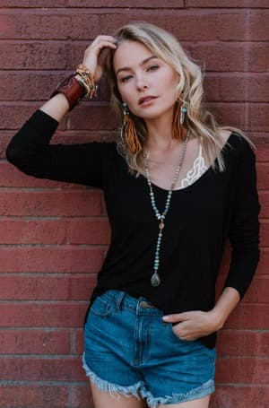 A blonde woman poses against a red brick wall wearing the Ryder Long Sleeve Bralette Tee - Black, paired with blue denim shorts, layered necklaces, large earrings, bracelets—one hand in her hair, the other in her pocket.