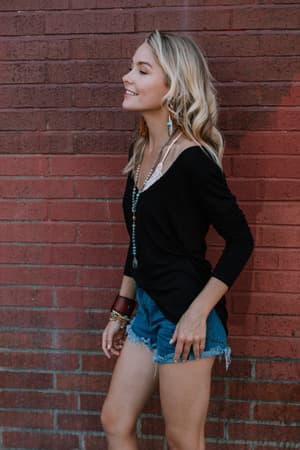 A woman with long blonde hair, smiling in profile against a red brick wall, wears the Ryder Long Sleeve Bralette Tee in black, paired with denim shorts, necklaces, and bracelets.