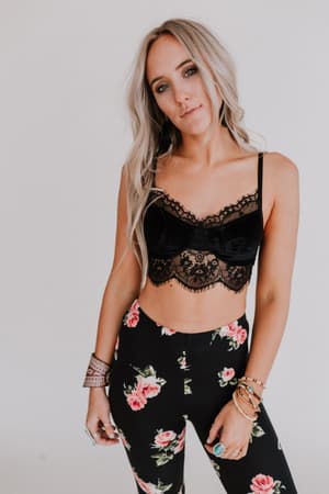 A woman with long, wavy blonde hair wears the Jessa Velvet and Lace Cami in black, paired with high-waisted black pants featuring a pink floral pattern. She accessorizes with bracelets and rings against a plain white background.