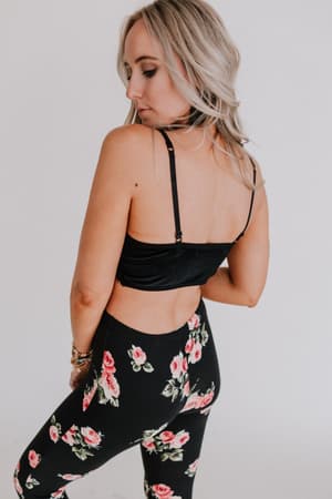 A woman with long blonde hair stands facing away, wearing the Jessa Velvet and Lace Cami in black with adjustable straps, paired with high-waisted black leggings featuring a pink floral pattern, set against a plain light background.