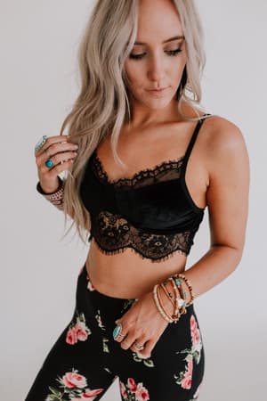 A person with long, wavy blonde hair wears the Jessa Velvet and Lace Cami - Black with adjustable straps, paired with black leggings featuring a pink floral pattern, accessorized with multiple rings, bracelets, and earrings, looking down thoughtfully.