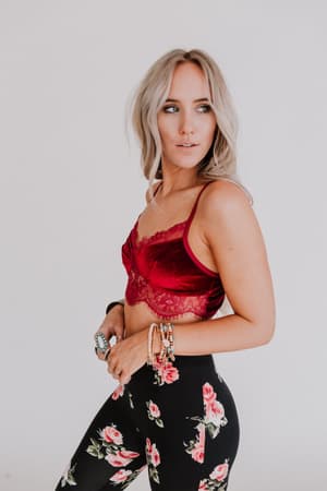 A woman with long blonde hair wears the Jessa Velvet and Lace Cami in burgundy, styled with high-waisted black leggings featuring a pink floral pattern, bracelets, and a turquoise ring, standing against a plain white background.