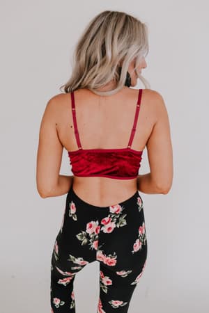 A woman with long, wavy blonde hair is seen from behind, wearing the Jessa Velvet and Lace Cami in burgundy with adjustable straps and black leggings featuring a pink floral pattern, standing against a plain light background.