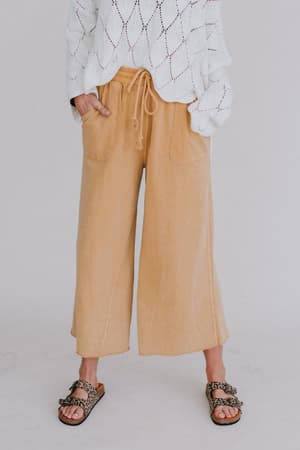 A person wearing the Relaxing Robin Wide Leg Pant in mustard, a white textured long-sleeve sweater, and leopard-print sandals stands against a plain light background. Only the lower half of their body is visible.