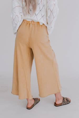 A person faces away, wearing Relaxing Robin Wide Leg Pant in mustard, a white textured sweater, and leopard print slide sandals against a plain, light backdrop.
