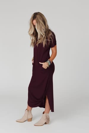 A woman with long, wavy blonde hair models the Call It Comfort Tee Dress in plum—a relaxed fit maxi dress with pockets—paired with beige fringe ankle boots and colorful bracelets against a plain light background.