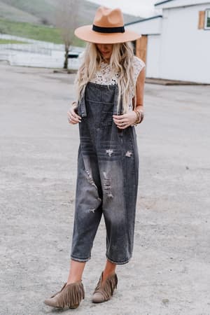 A woman in a tan hat, white lace top, and High Hopes Slouchy Denim Overalls - Black stands outdoors on concrete. She has long blonde hair and wears tan fringe ankle boots.