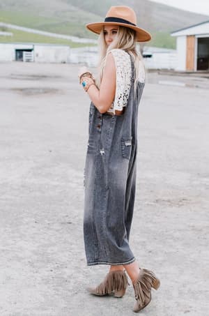 A woman in a tan hat, white lace top, and the High Hopes Slouchy Denim Overalls in black stands outdoors on a paved surface in tan fringed ankle boots, glancing over her shoulder.