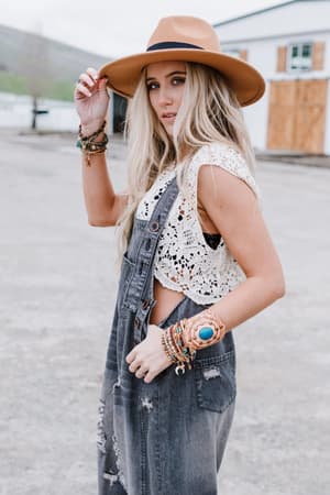 A woman wearing the High Hopes Slouchy Denim Overalls in black, a tan wide-brim hat, and a white crocheted top stands outdoors. She has long blonde hair, layered bracelets, and gazes at the camera while adjusting her hat.