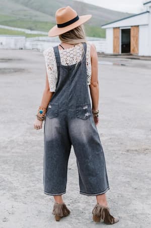 A woman with long blonde hair stands outdoors facing away, wearing a tan hat, white lace top, and the High Hopes Slouchy Denim Overalls in black. She pairs them with brown fringed ankle boots and multiple bracelets on both wrists.