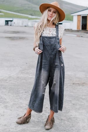 A woman in a tan hat, white lace top, High Hopes Slouchy Denim Overalls - Black, and taupe fringe shoes stands outdoors on concrete with a rural backdrop.